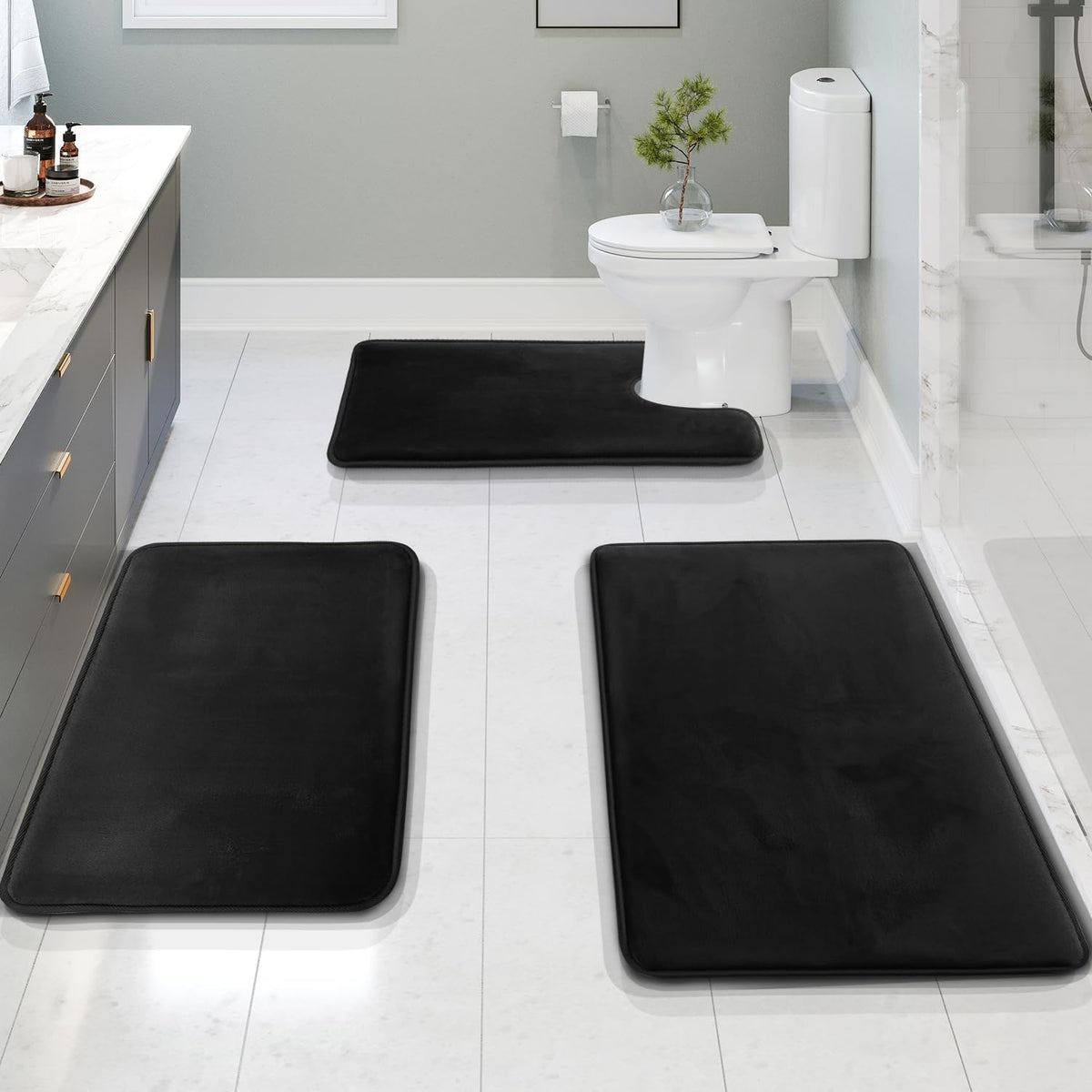 Low-angle bathroom view showing three black memory-foam rugs laid on glossy white tile toward a toilet and vanity.