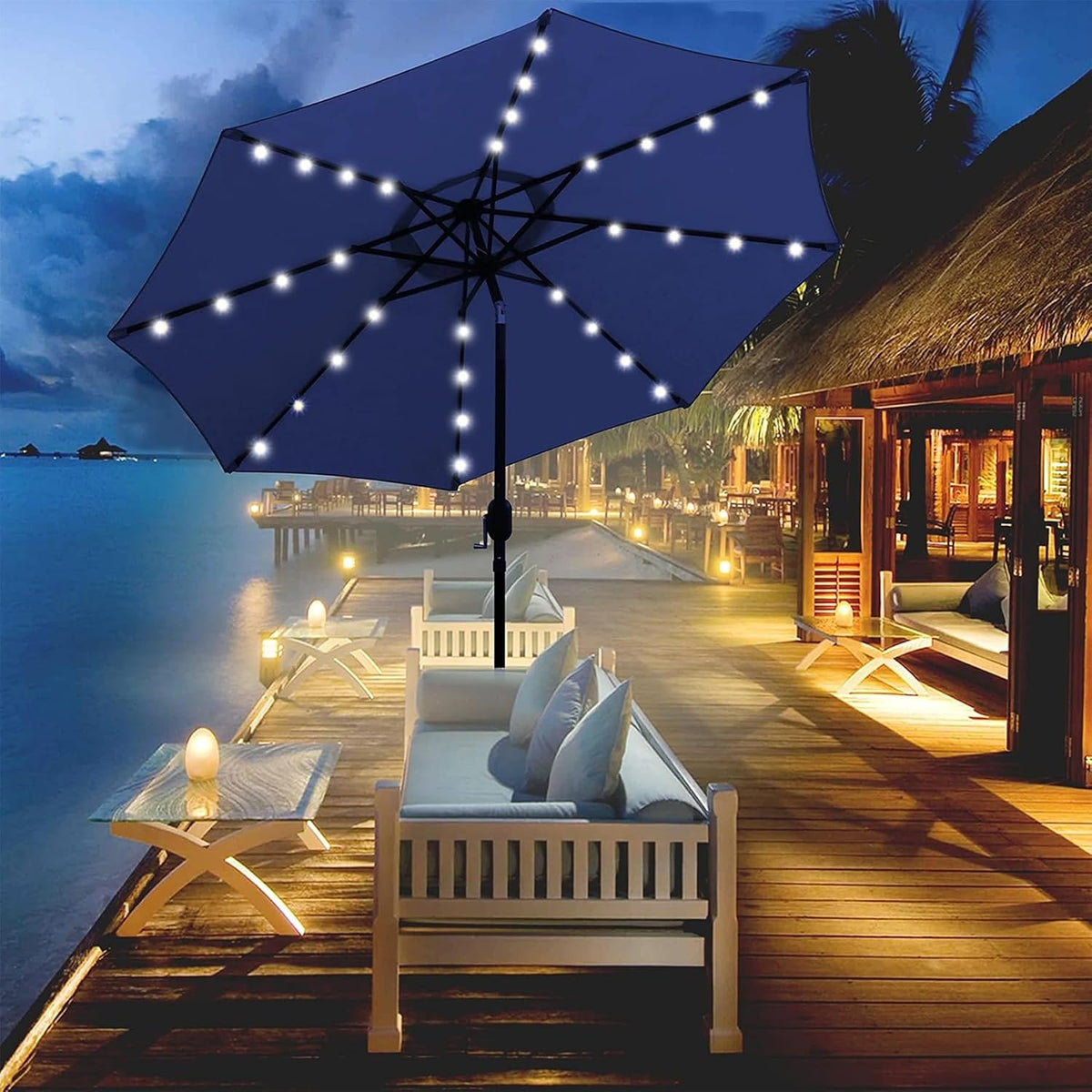 Navy blue LED umbrella glowing over outdoor seating beside a waterfront at dusk