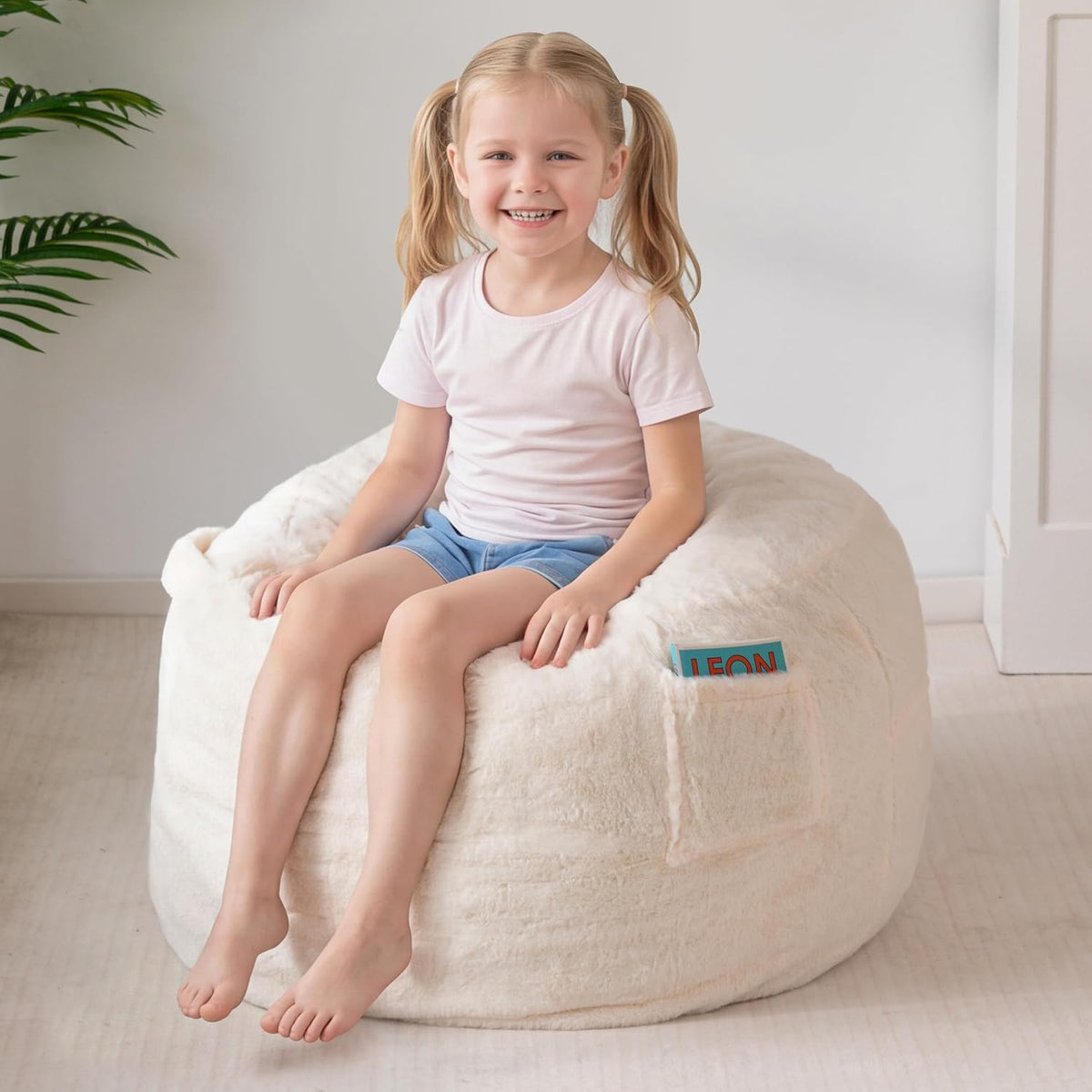 Young girl smiling while seated on a cream-colored plush bean bag with a side pocket holding a book.