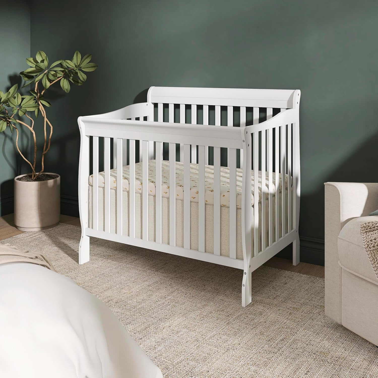 White crib placed in a styled nursery corner with plant, rug, and sofa visible