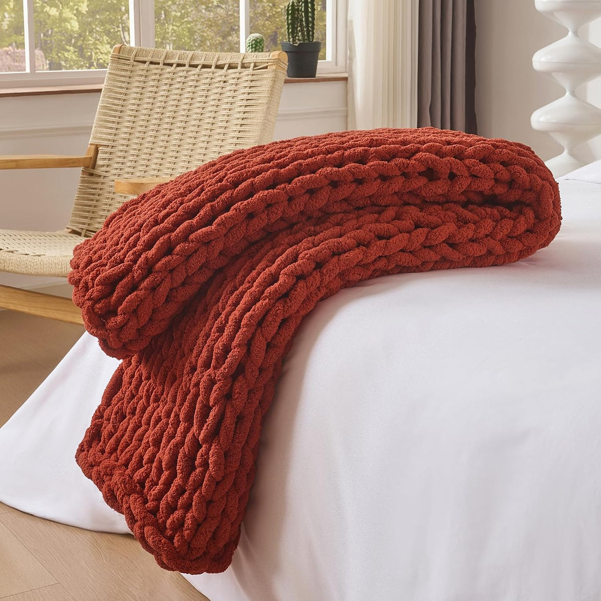 Folded rust-colored chunky knit throw resting on the edge of a bed near a woven chair