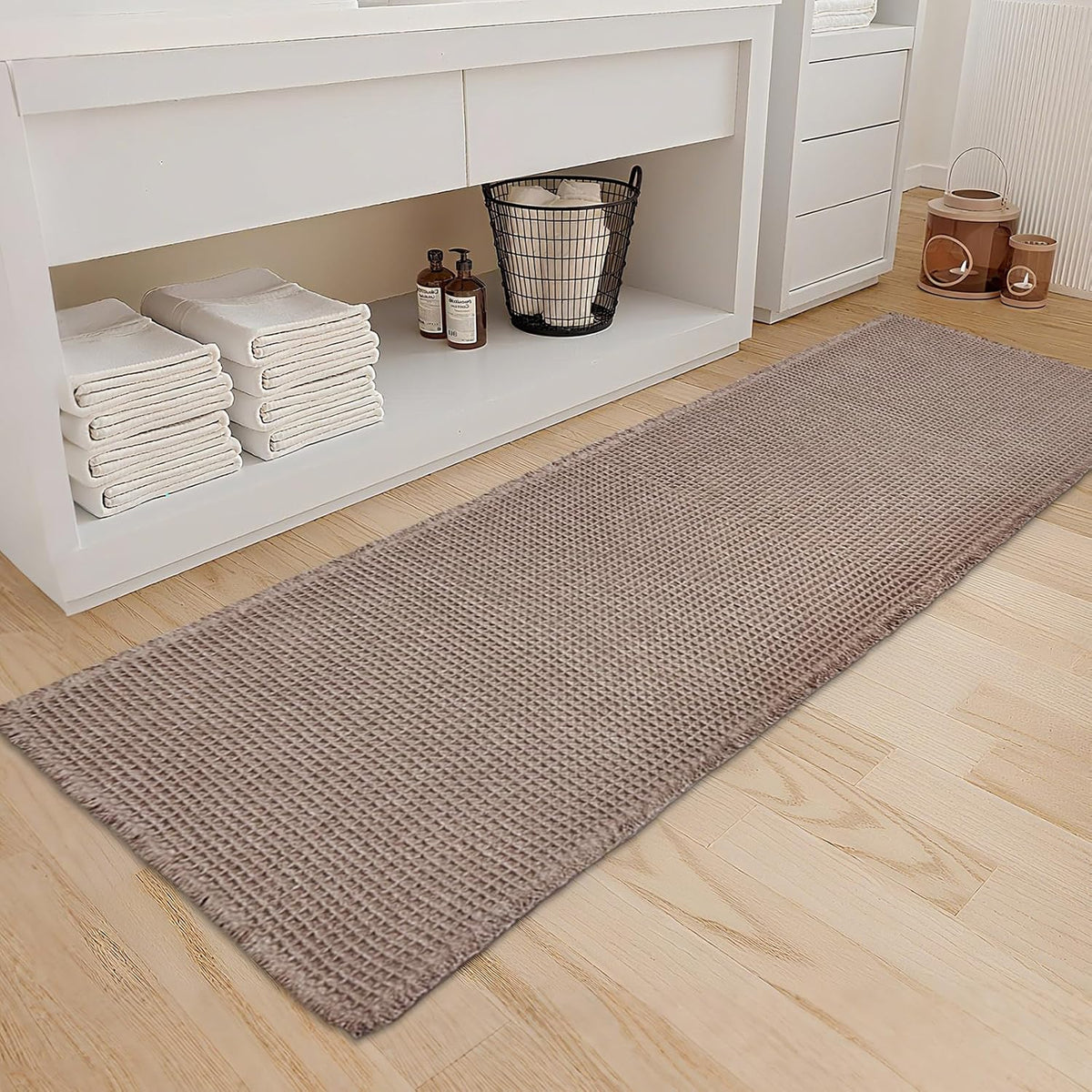 Close angled view of a tan waffle runner on a pale wood floor beside white bathroom storage and stacked towels.