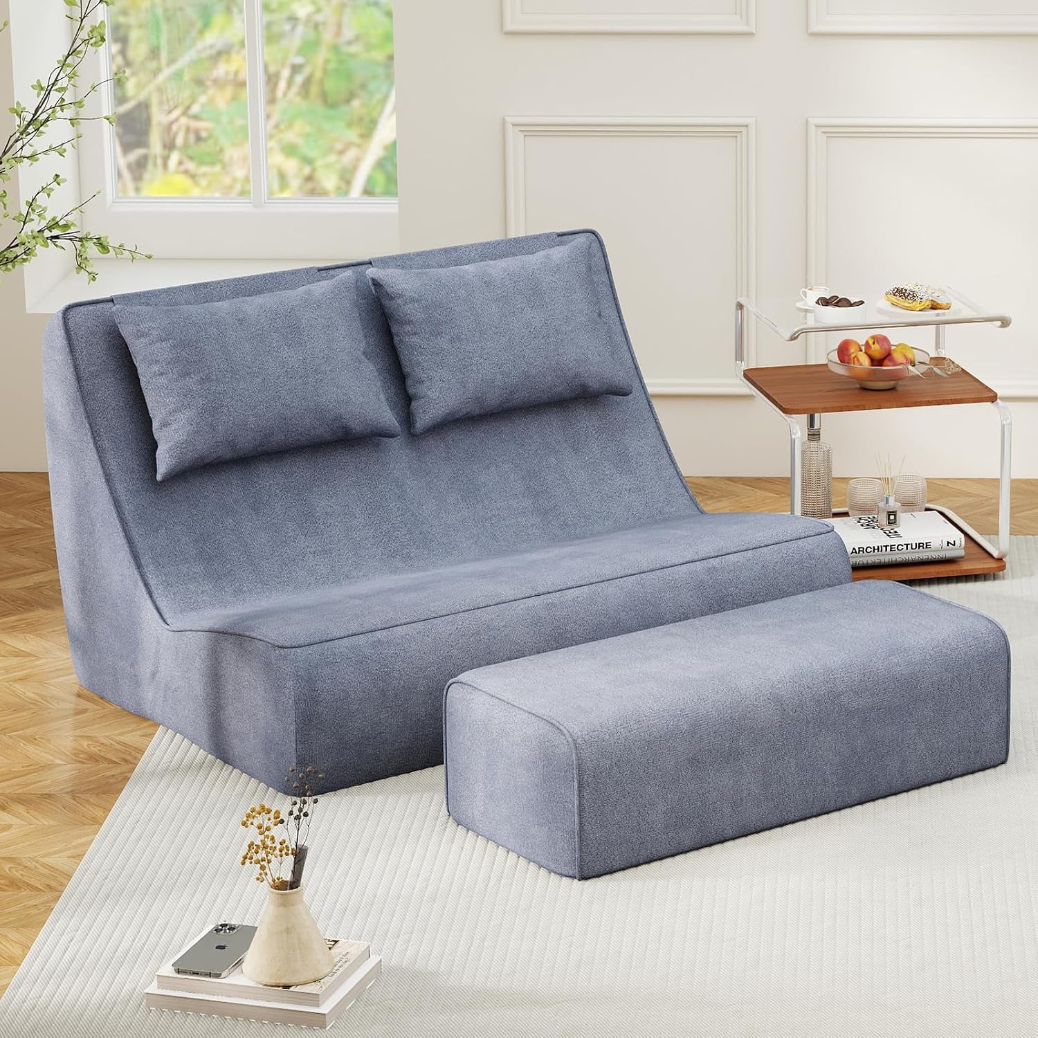 Blue chaise lounge with two loose pillows and matching ottoman on wood parquet flooring near a window.