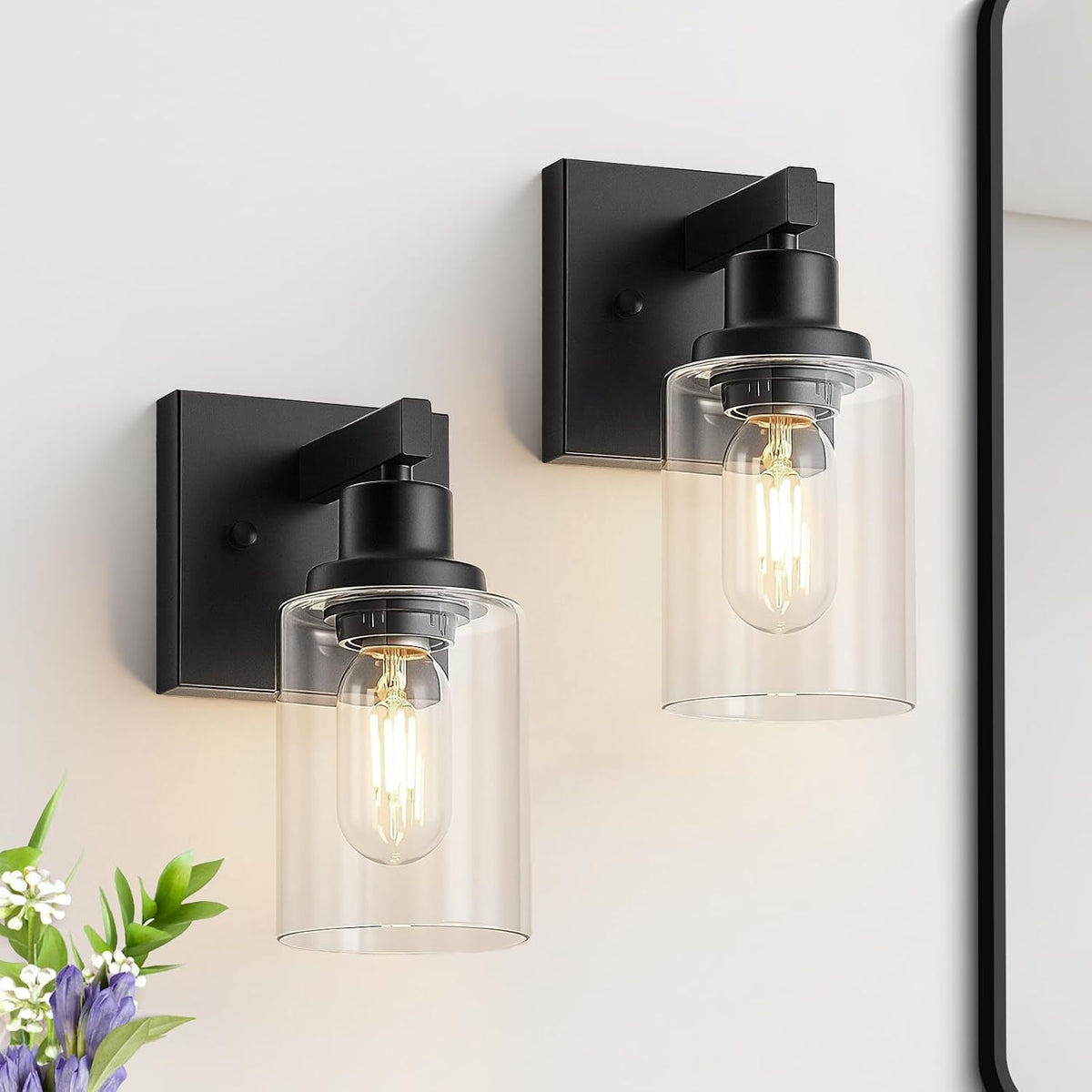 Pair of single-light matte black wall sconces with clear cylindrical shades shown next to a mirror edge.