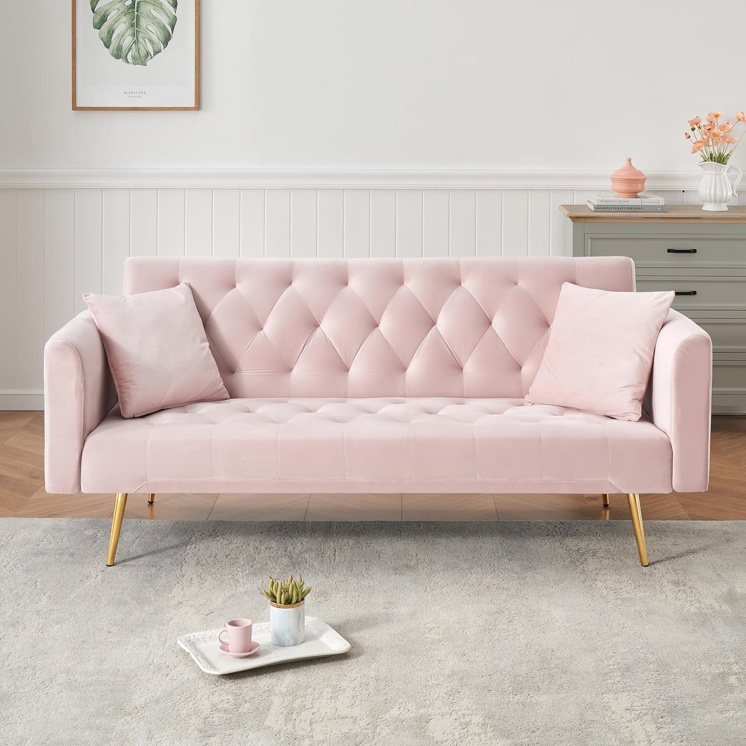 Straight-on view of a pale pink tufted velvet sofa with gold legs, tray and cup on a rug in front.