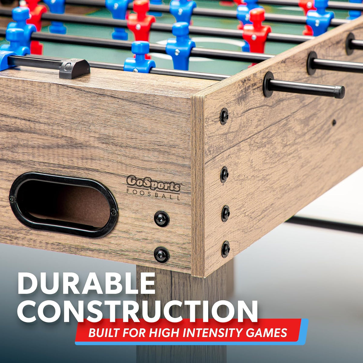 Close-up of a wood-finish foosball table corner showing ball retrieval port, screw details, and textured edge