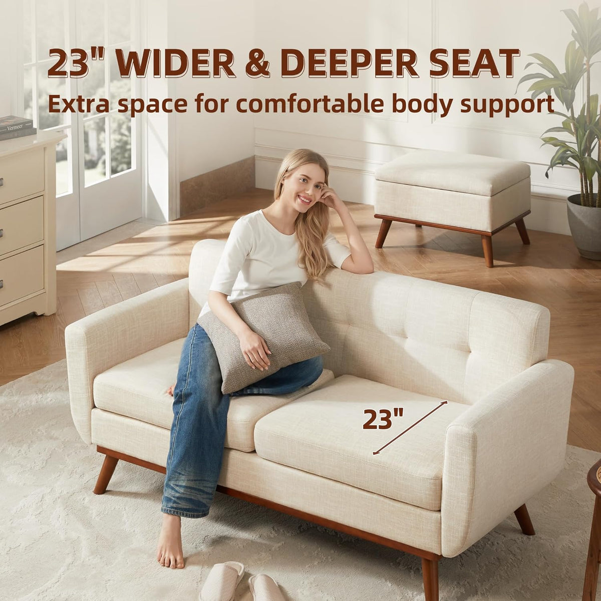 Angled lifestyle shot of a beige loveseat with a woman seated, matching ottoman and sunlit room background.