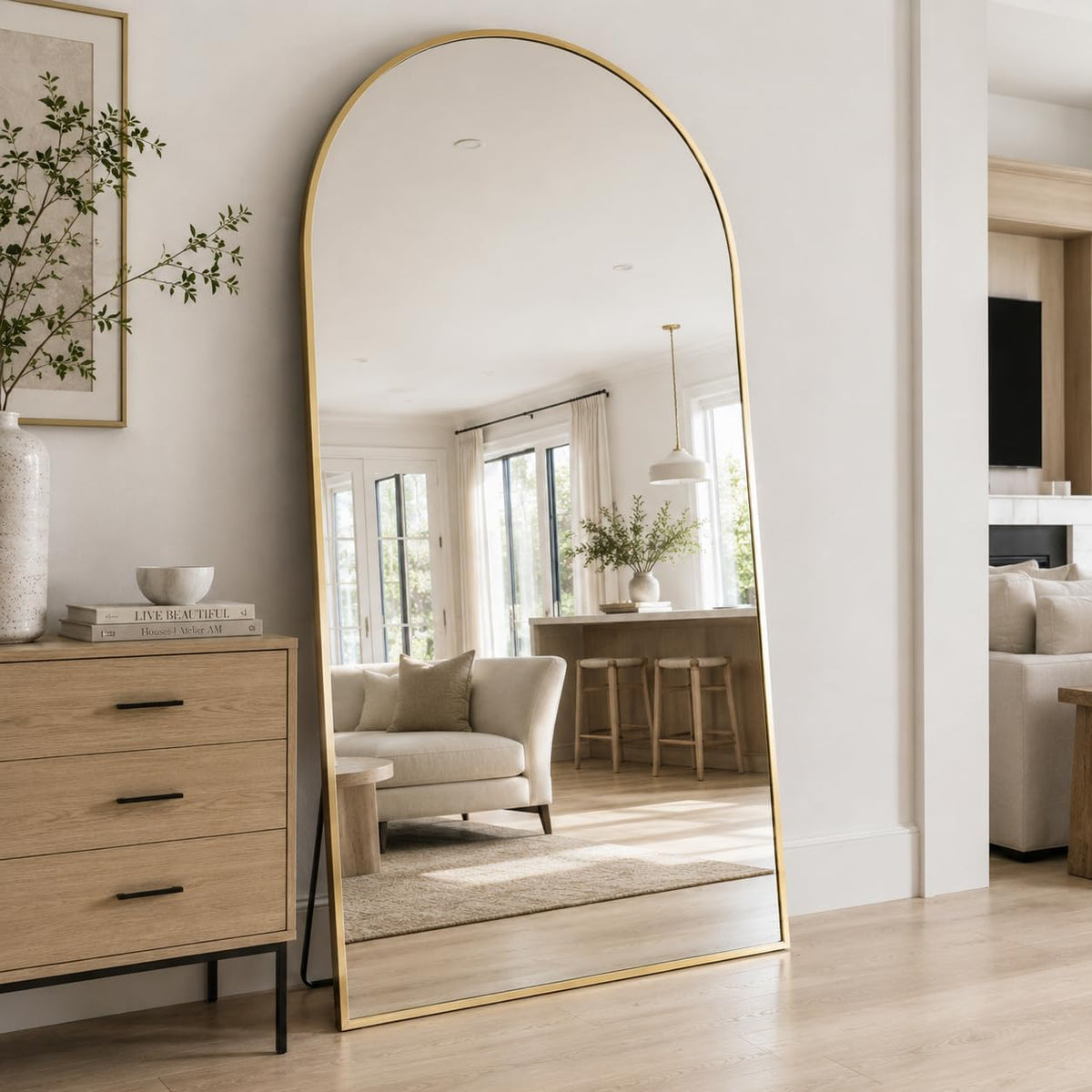 Tall arched mirror with a gold frame leaning on the floor beside a wood dresser and neutral living room.
