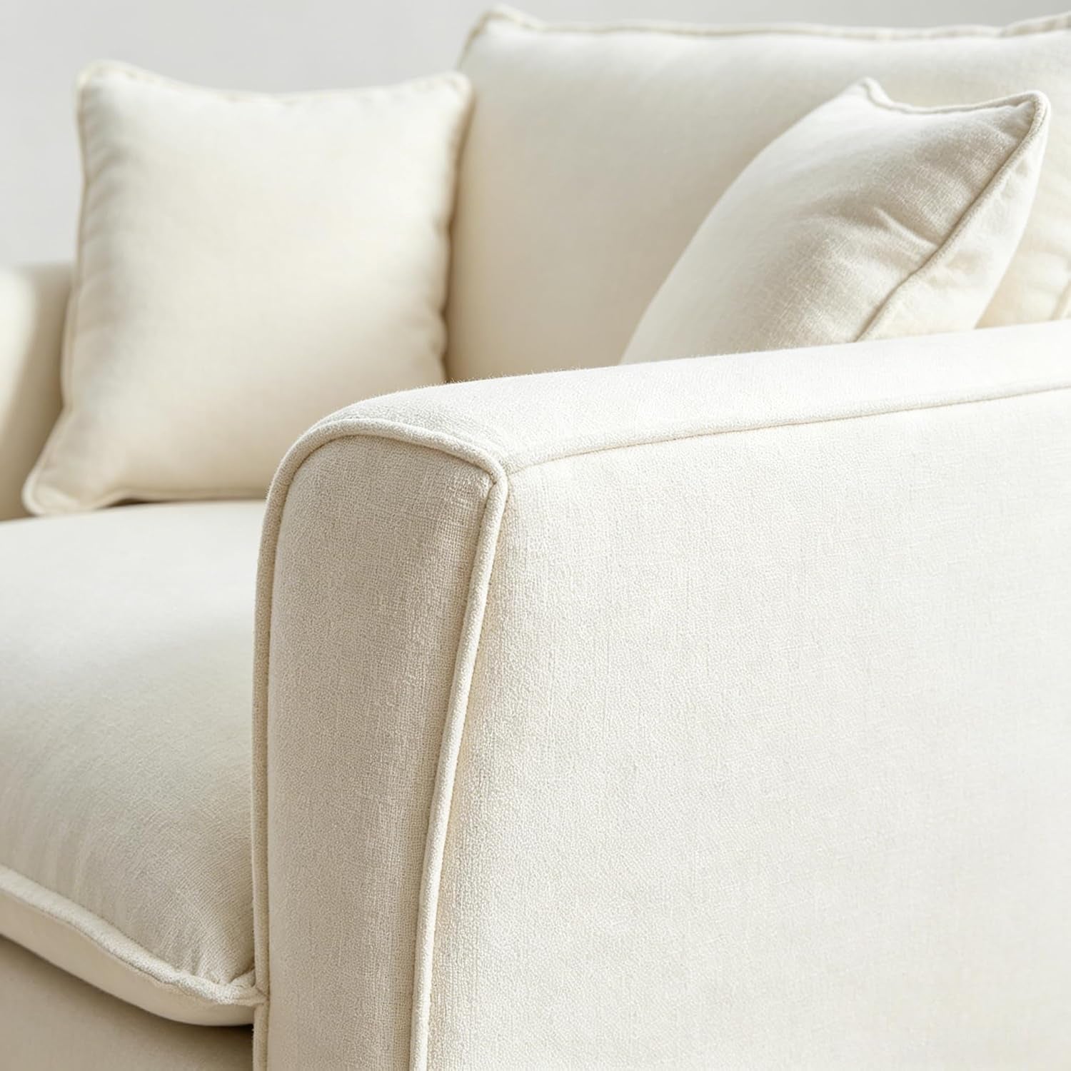 Close-up detail of the chair's arm and back showing cream textured chenille fabric and piped seam with two pillows blurred