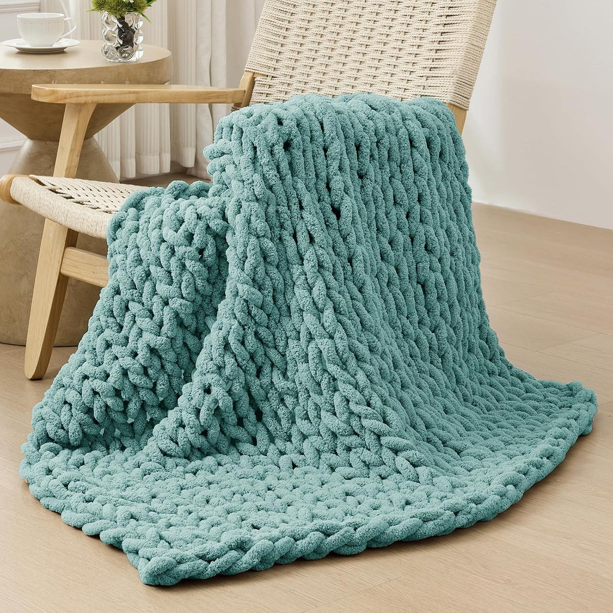 Teal chunky knit throw draped over a woven armchair, pooling onto a light wood floor.