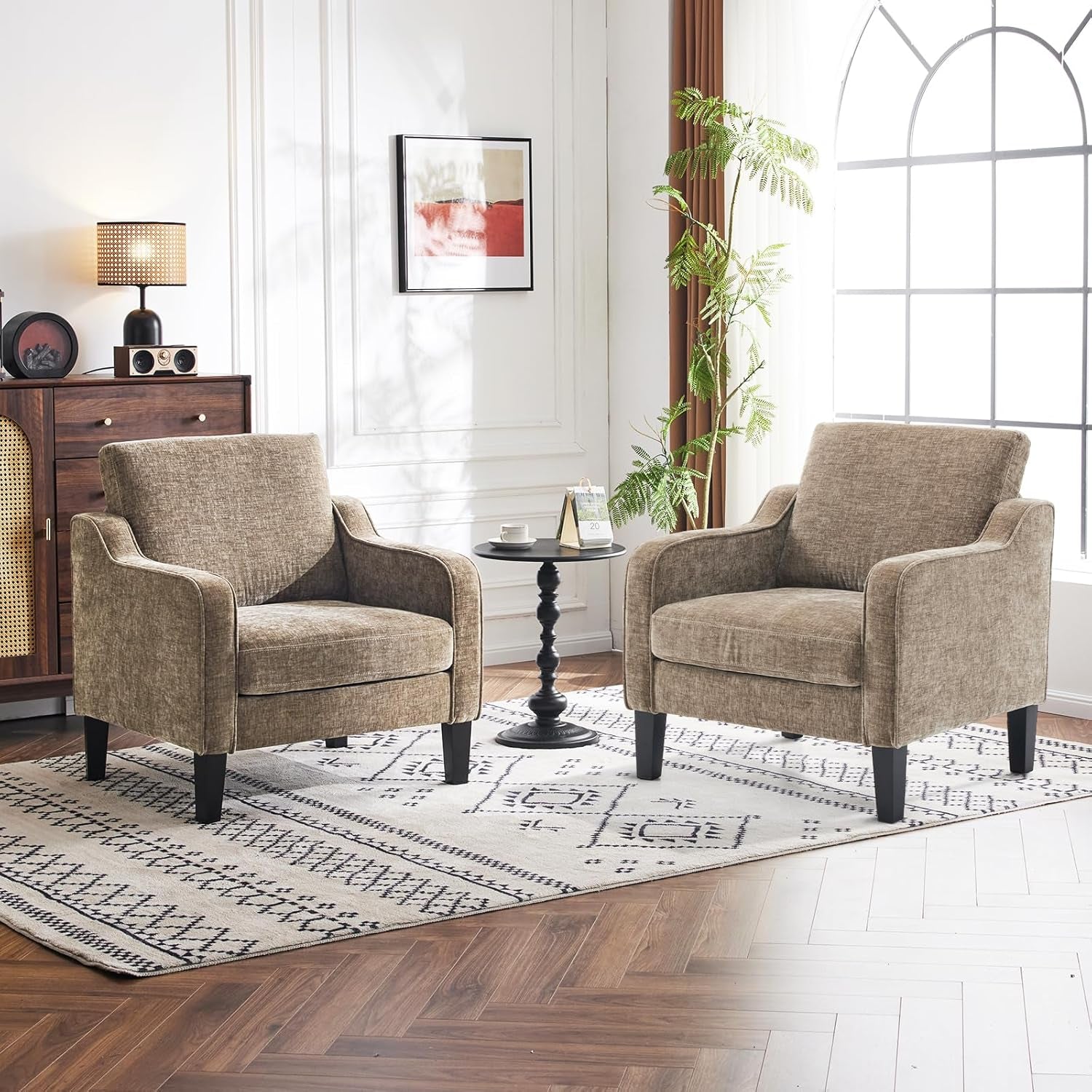 Two tan-brown upholstered accent chairs arranged around a black pedestal side table on a patterned rug.