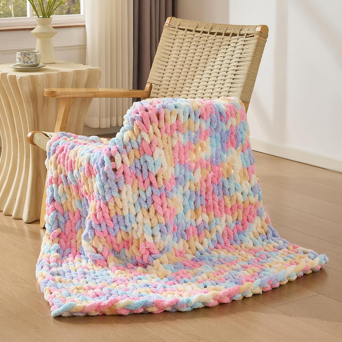 Pastel multicolored chunky knit throw draped over a woven wood chair, pooling on the floor.