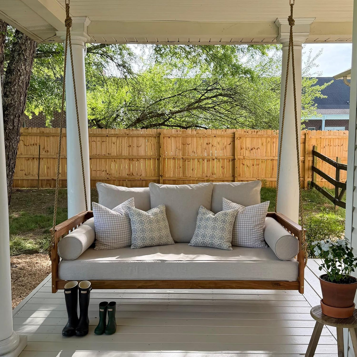 Acacia daybed porch swing in oil finish with light cushions, front view on a covered porch with fence background
