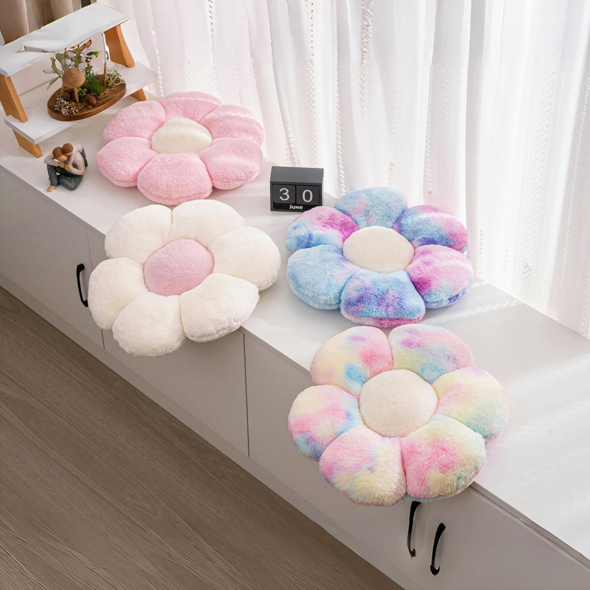 Three daisy throw pillows in pink, white and blue tie-dye displayed on a white cabinet by a sheer curtain