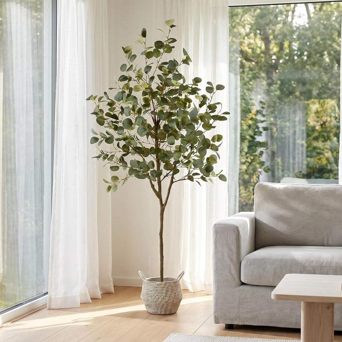 6 ft artificial eucalyptus tree in a woven basket next to a sofa by floor-to-ceiling windows in a sunlit room