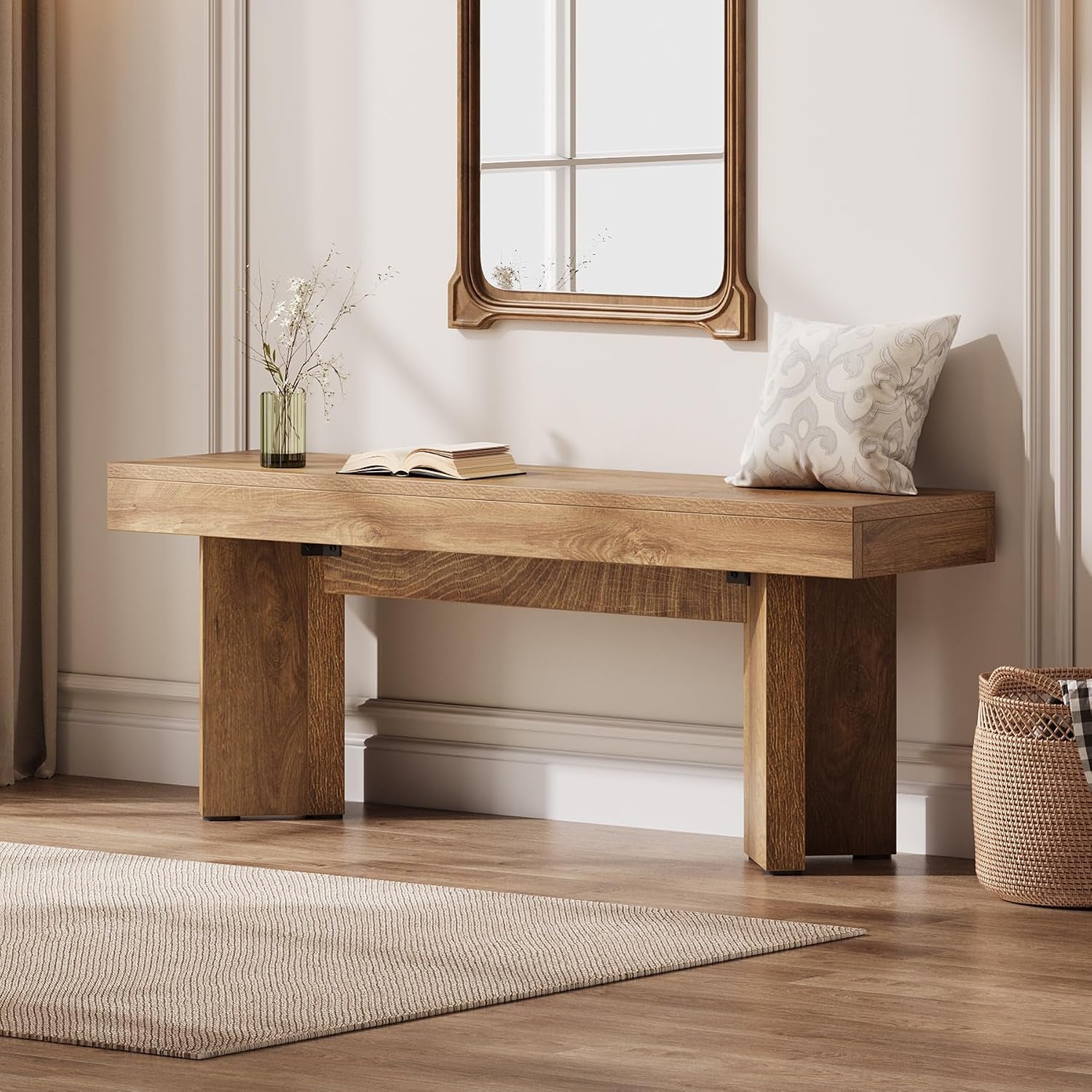 Rustic brown bench against a paneled wall under a mirror with a patterned pillow, vase and open book on top.