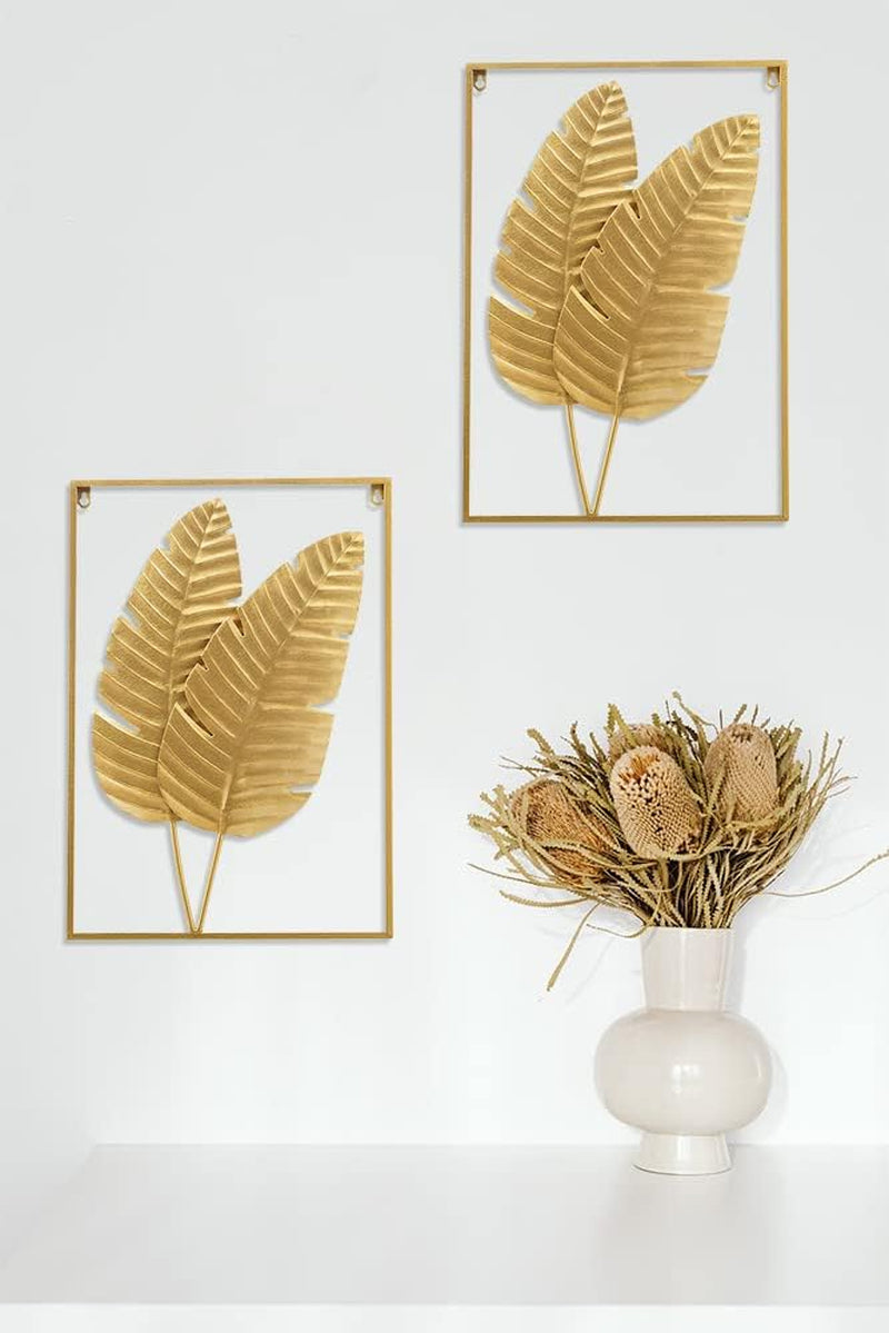Two gold leaf frames hung on a white wall above a white surface with a chunky ceramic vase holding dried protea stems.