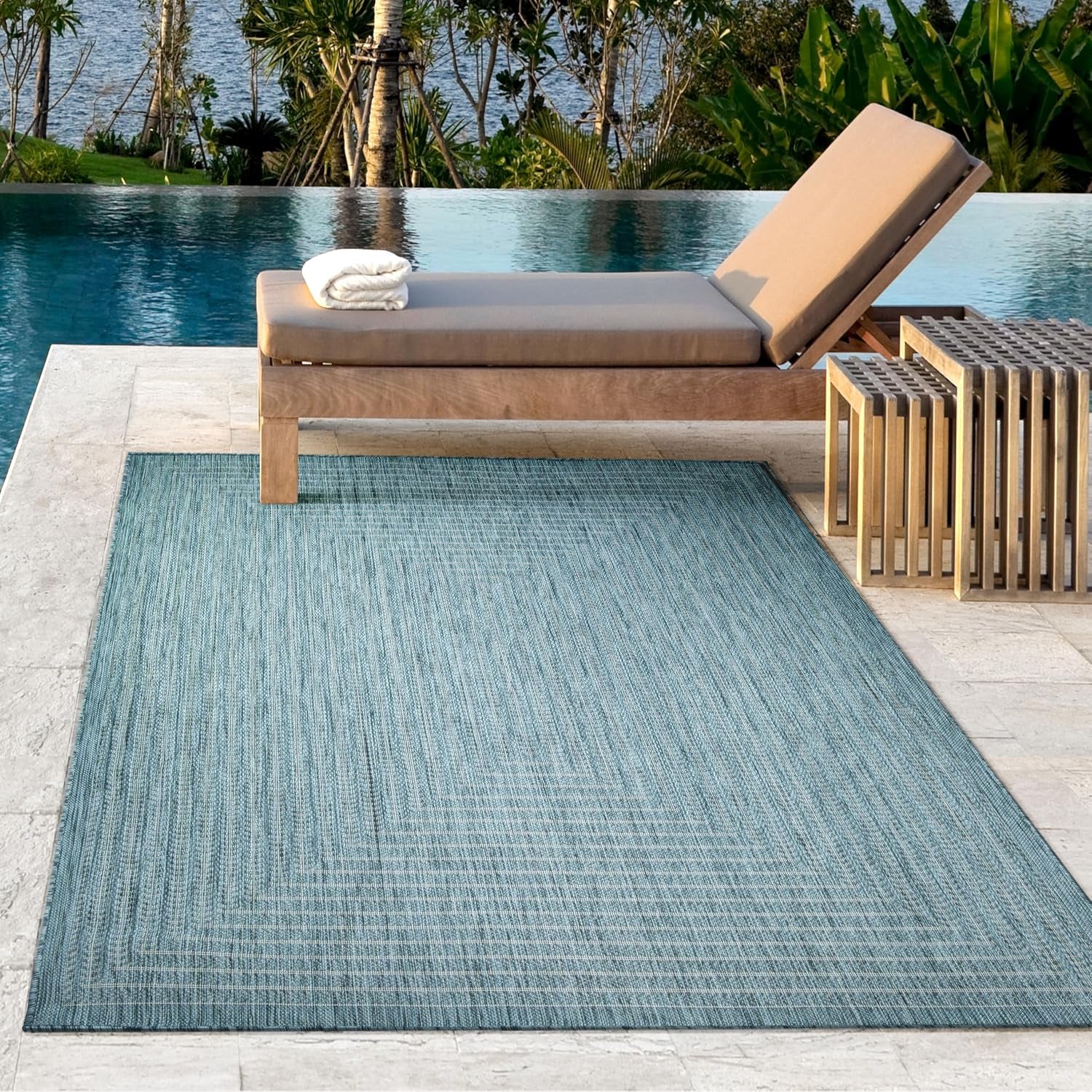 Aqua outdoor rug placed beside a pool under a wooden chaise lounge and small slatted side tables.