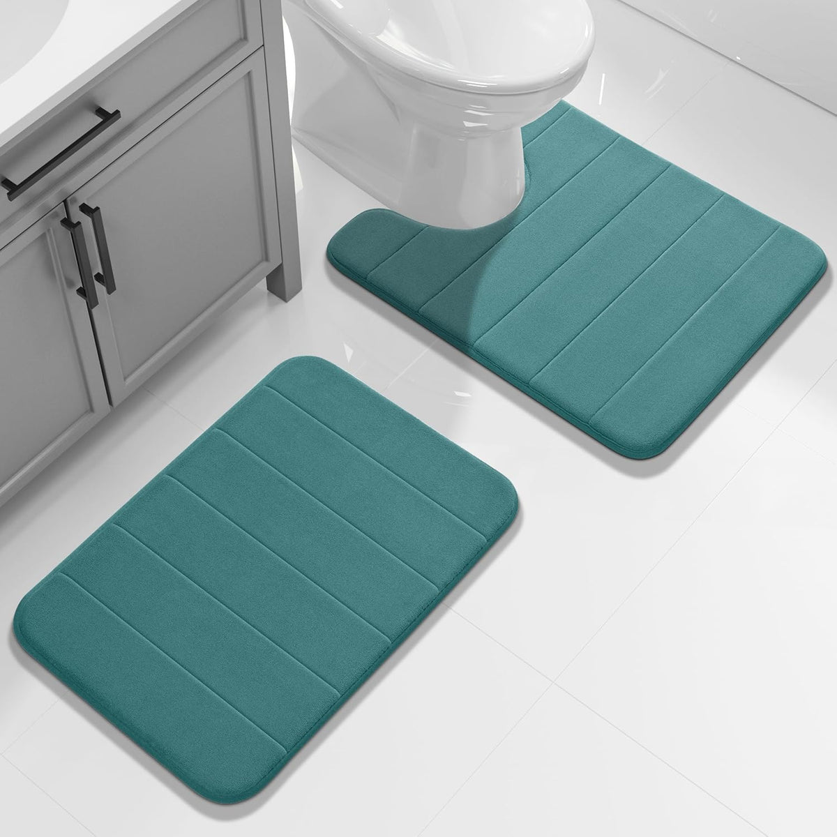 Two rectangular bathroom mats in teal placed on a glossy white tile floor near a vanity and toilet.