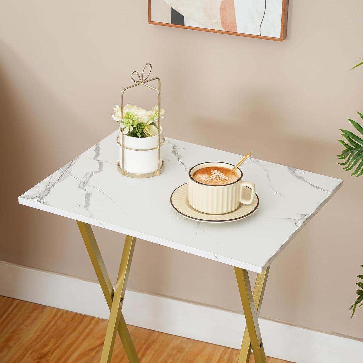 Marble-pattern folding tray table with gold legs holding a cup and small plant against a beige wall