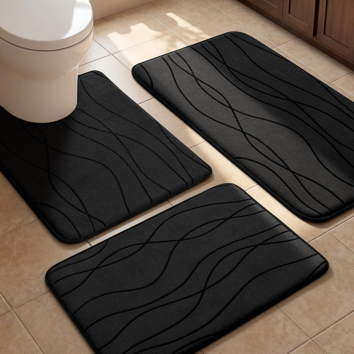 Three dark-gray wavy-pattern bathroom mats arranged on tiled floor near a toilet and wooden cabinet.