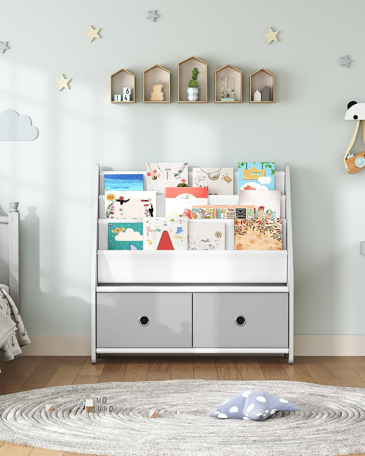 White four-tier kids bookshelf with front-facing books and two gray fabric storage bins in a pastel nursery.