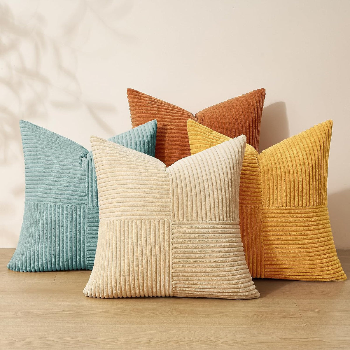 Five square corduroy throw pillows in teal, cream, rust and mustard arranged against a pale wall.