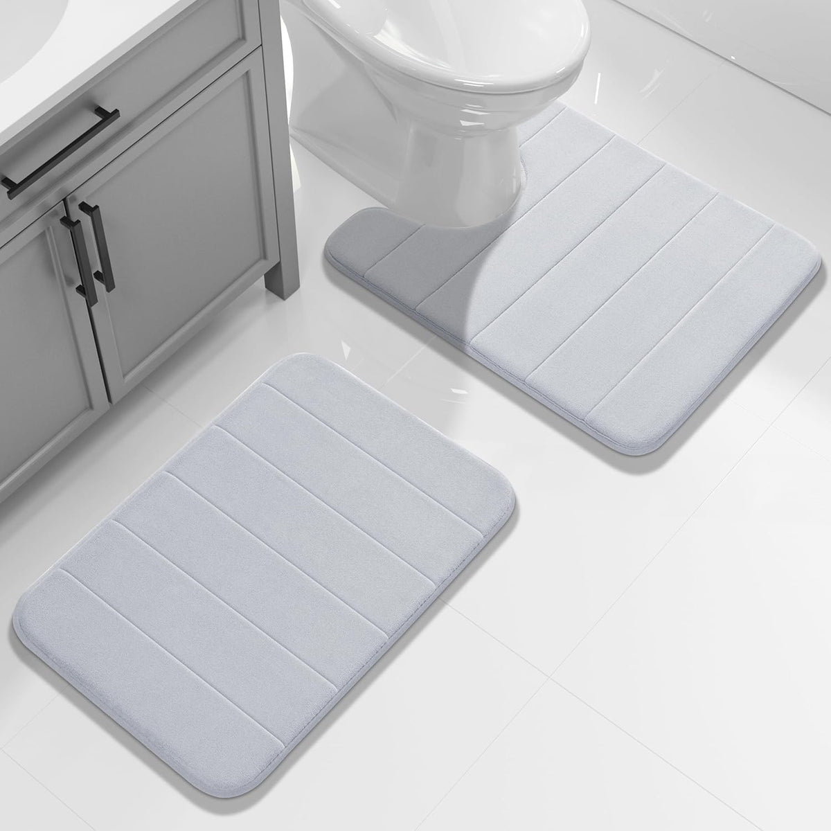 Pair of light gray rectangular bathroom rugs placed by a sink cabinet and toilet on white tile.