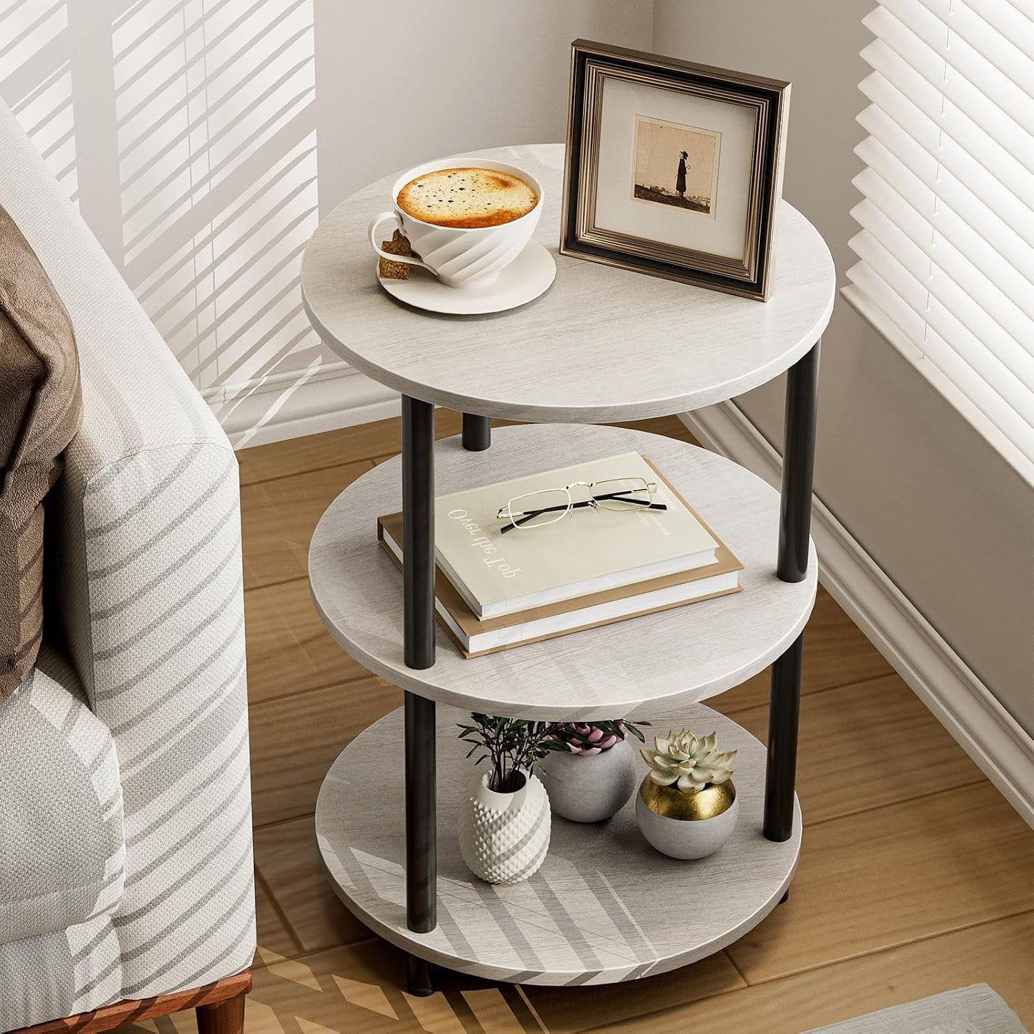 Three-tier round side table in light gray beside a sofa with a cup, framed photo and small plants.
