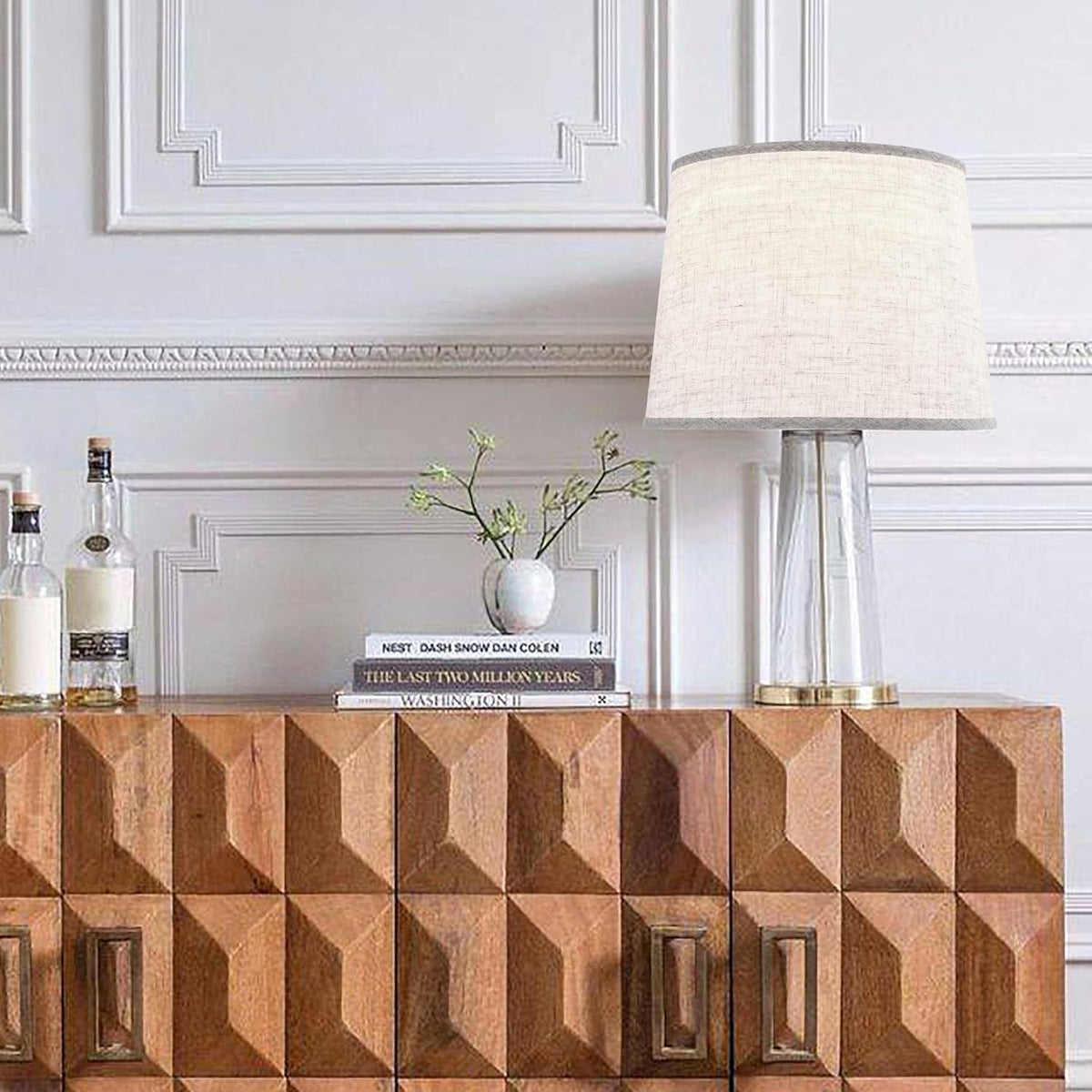 Table lamp with an oatmeal linen drum shade on a geometric wood sideboard beside books, a small vase, and bottles.