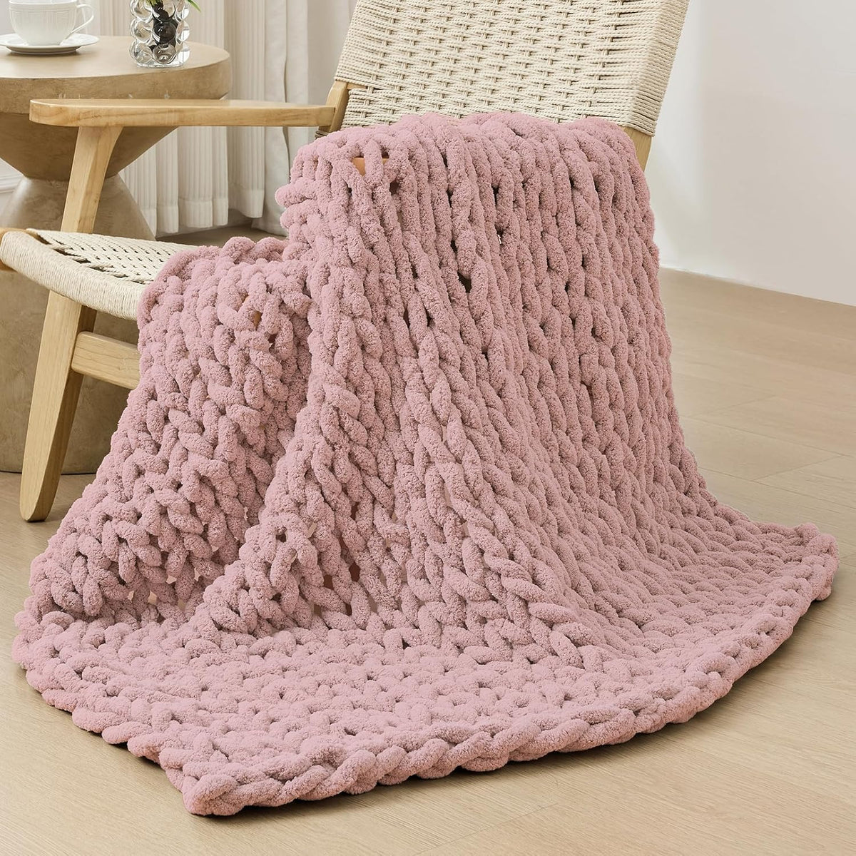 Dusty rose chunky knit throw casually draped over a woven chair with the blanket spread on the floor.