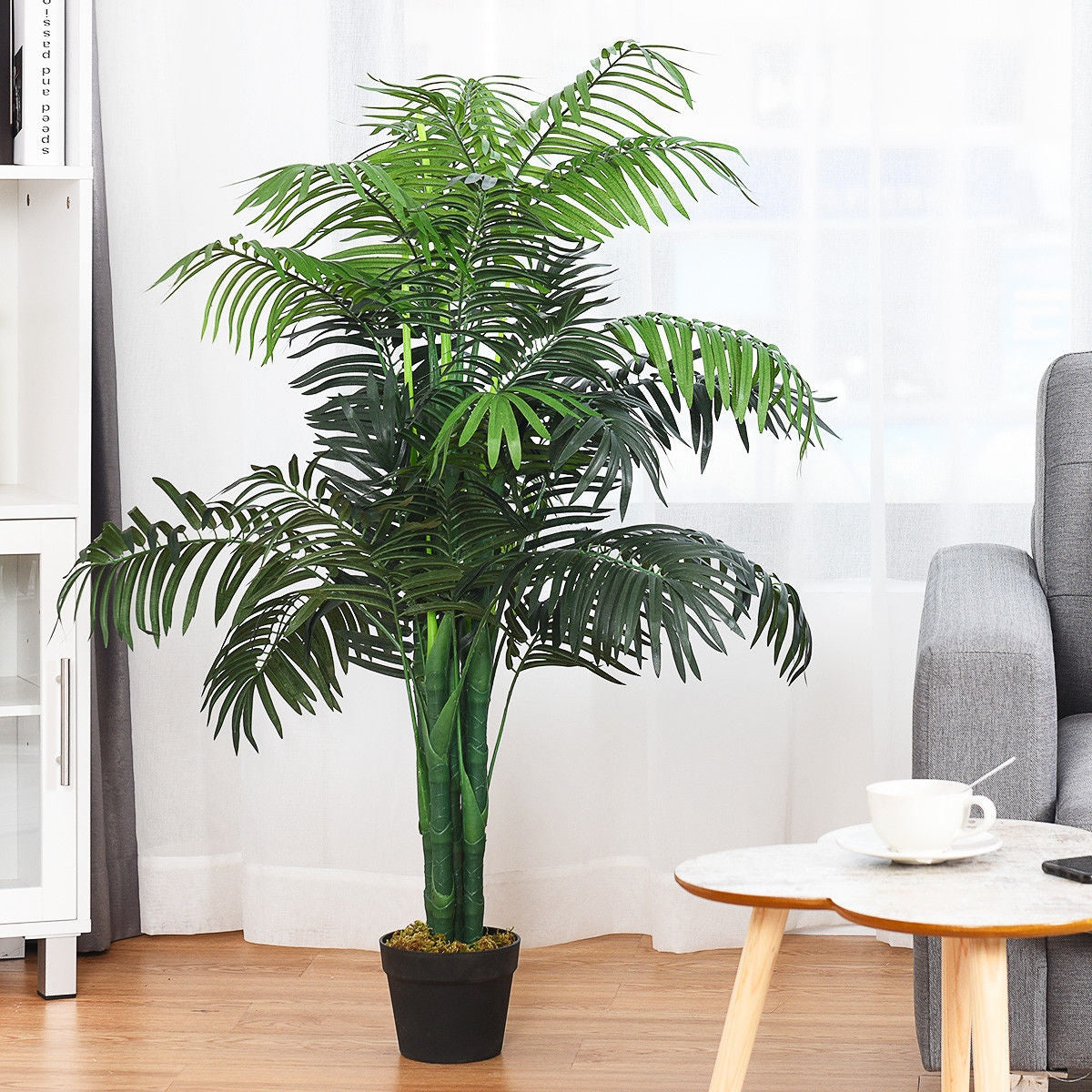 Potted artificial palm stands beside a white cabinet and gray chair near a bright window.