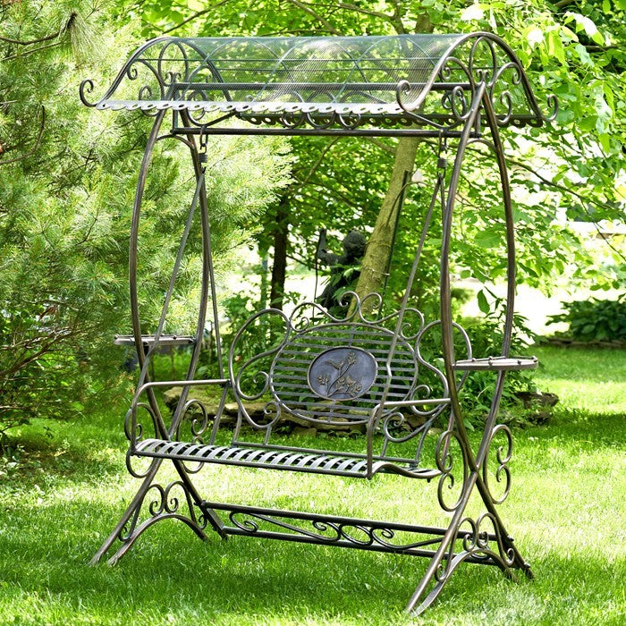 Ornate metal swing bench with a curved mesh canopy stands on a lawn beneath leafy trees.