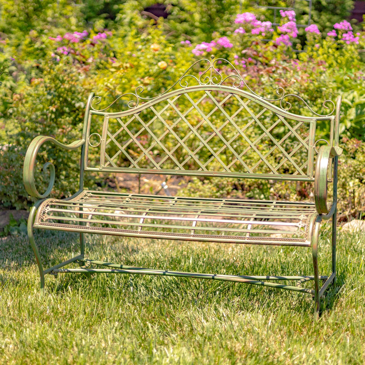 Antique green iron garden bench shown front-facing on grass among flowering shrubs