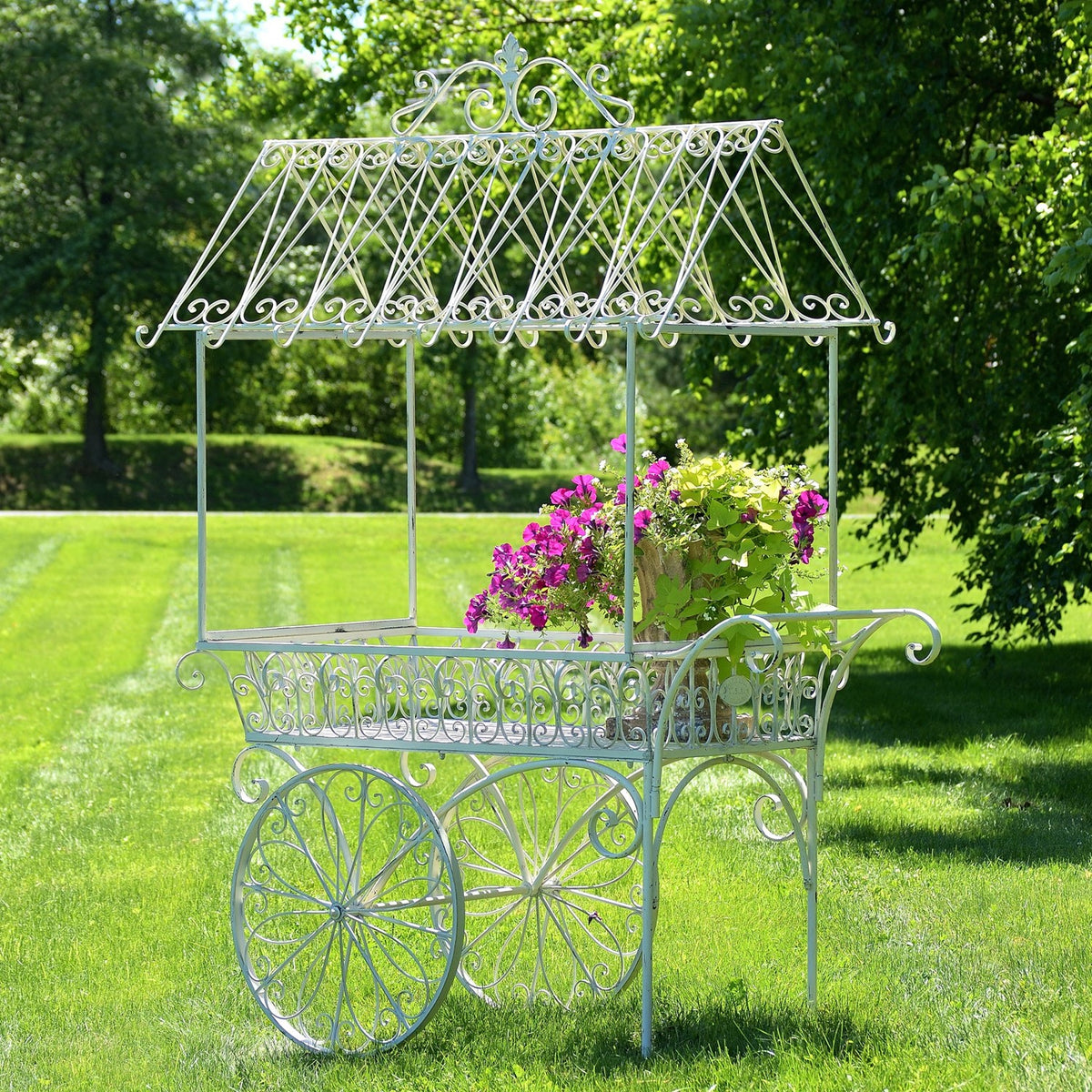 Ornate white iron flower cart with a peaked lattice canopy and pink flowers on a sunny lawn.