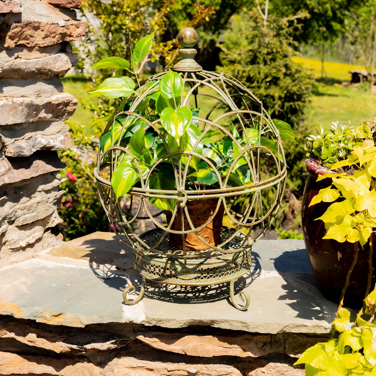 Green-toned wire globe plant stand holding a leafy plant beside a stone wall