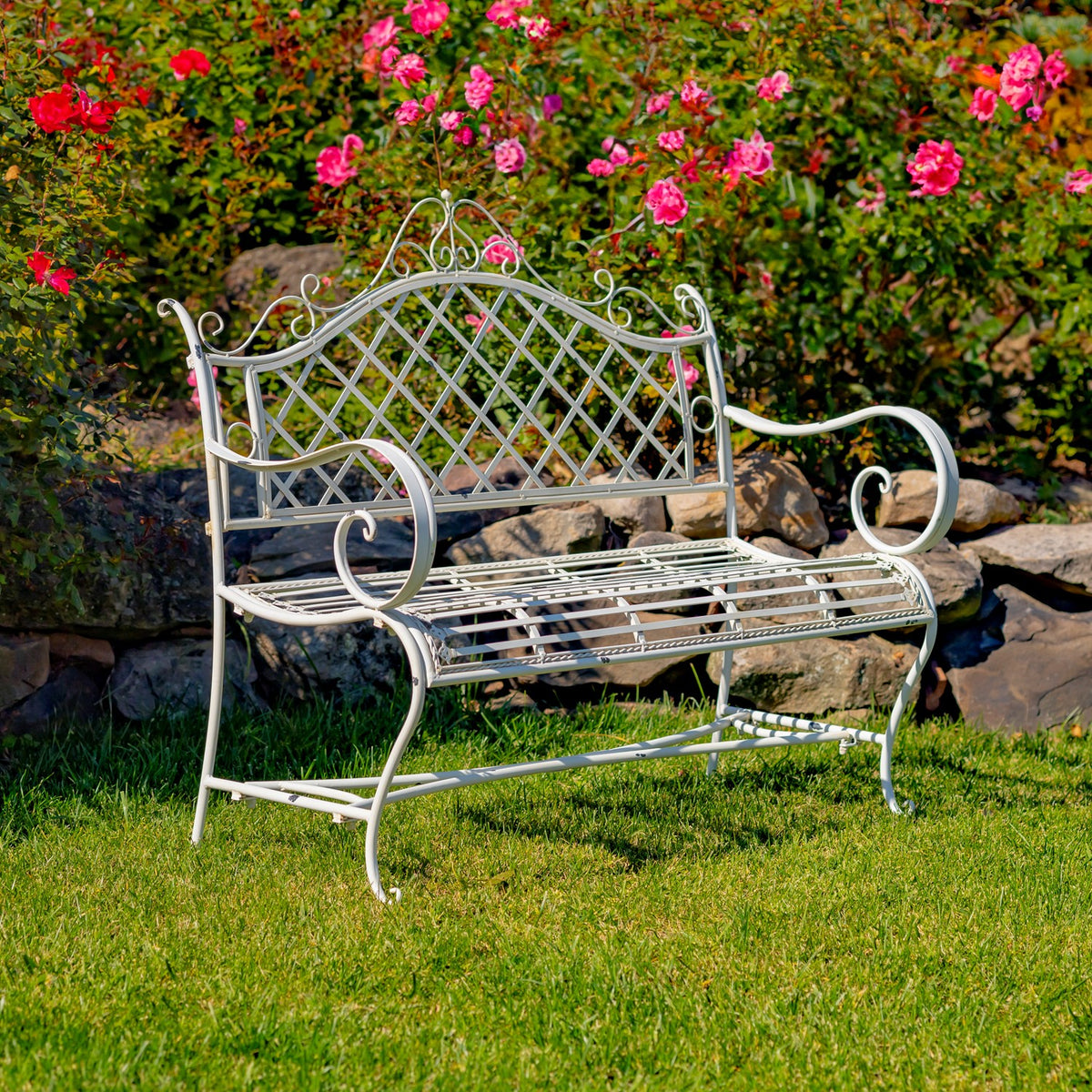 Antique white lattice-back iron garden bench on grass beside flowers and a stone border