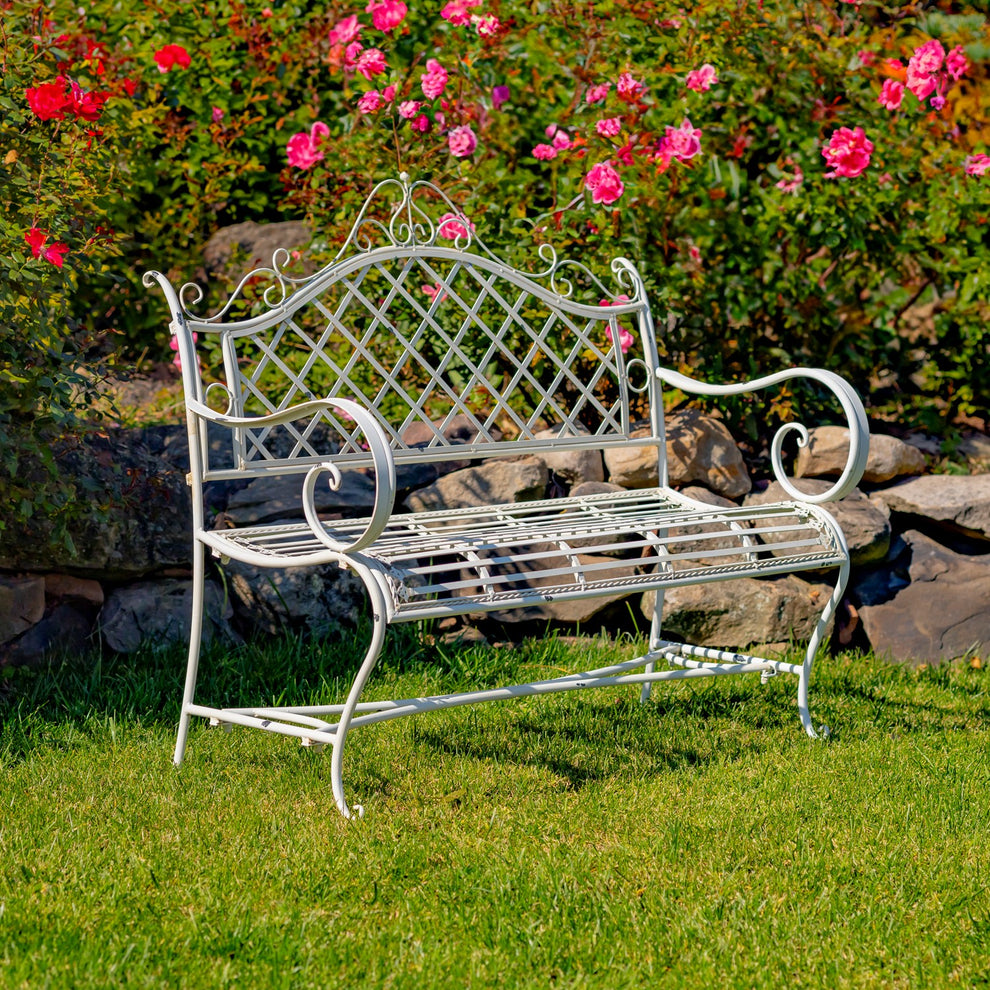 Antique white lattice-back iron garden bench on grass beside flowers and a stone border