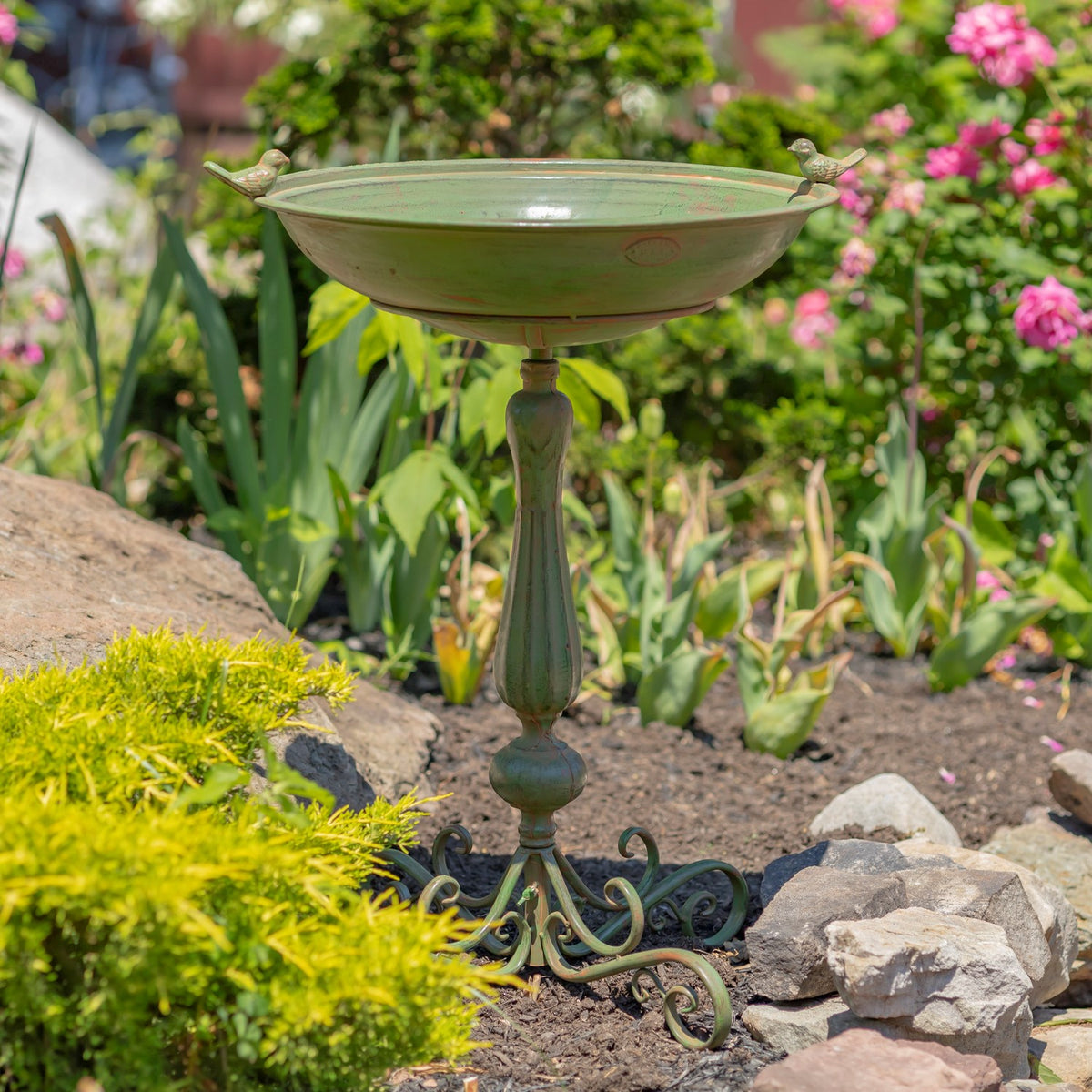 Green pedestal birdbath with ornate curled feet and small birds on the rim, set among garden flowers and stones.