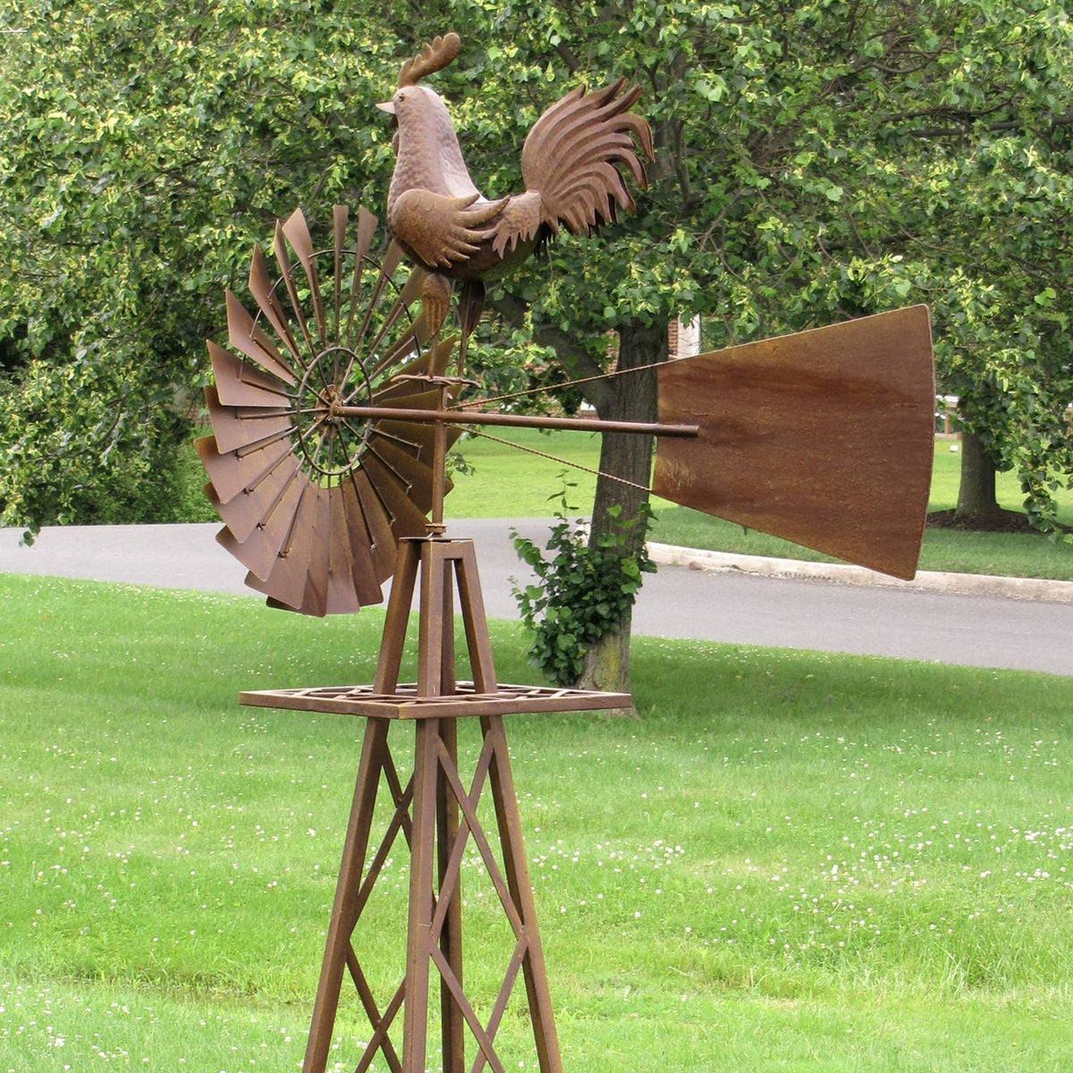 Rust-colored windmill stand with a rooster ornament, shown among trees and lawn.