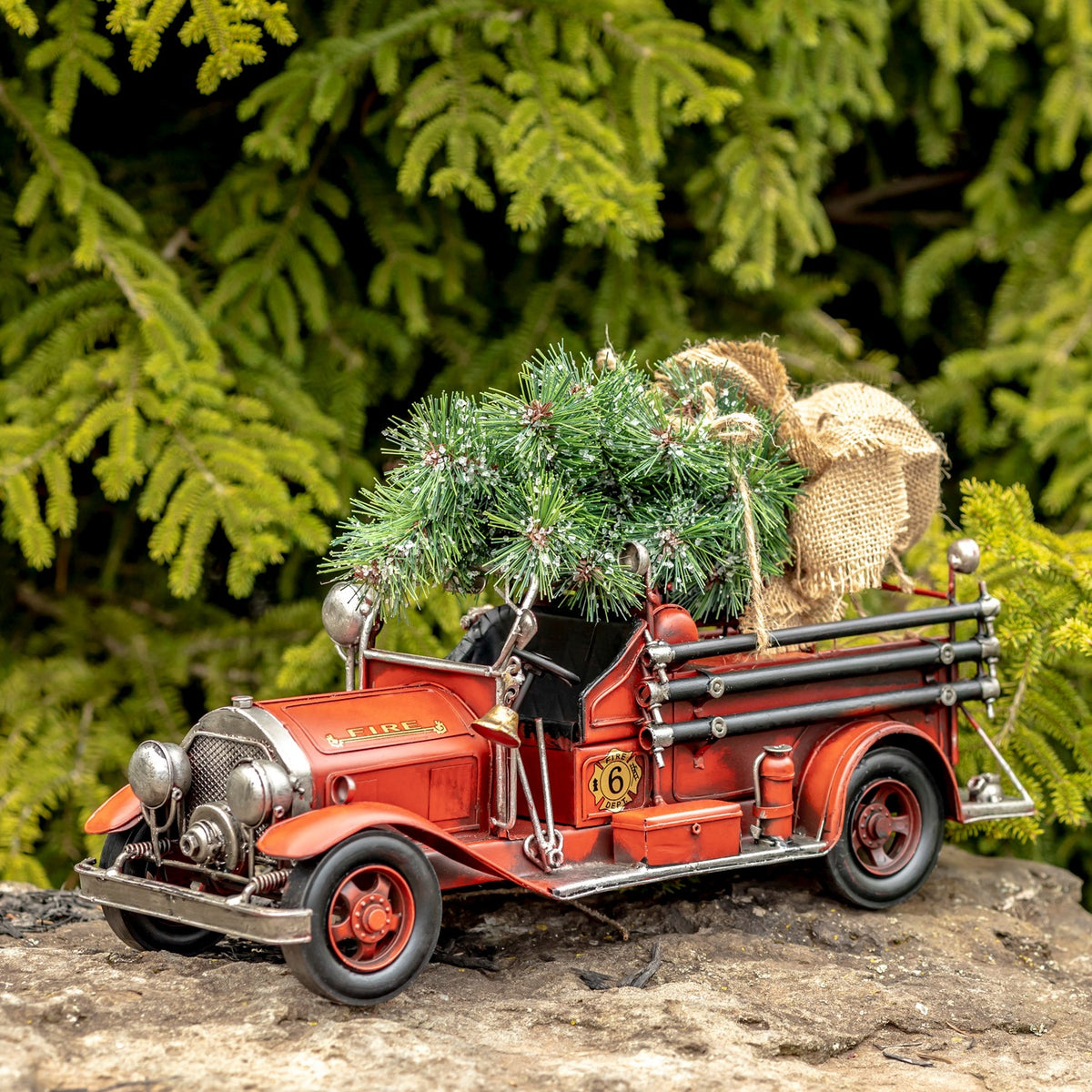 Red vintage-style fire truck carrying a small evergreen tree and burlap sack, shown from the front side outdoors.