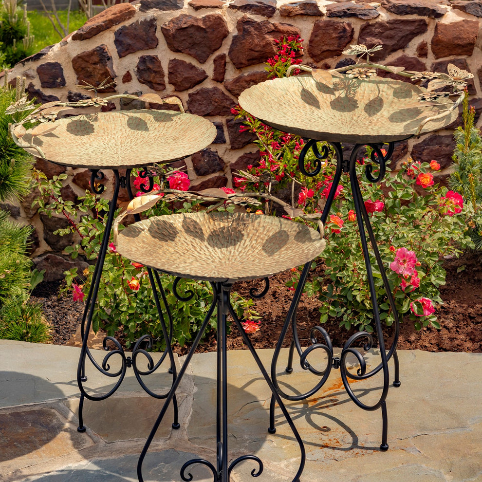 Three gold-toned birdbaths with black scrollwork stands arranged among garden flowers.