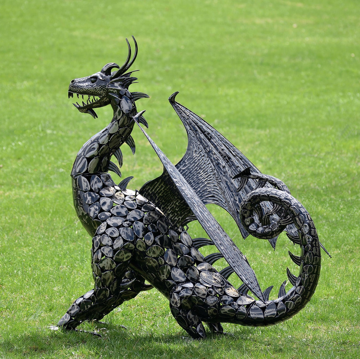 Black metal dragon statue standing on grass, with spread wings and a tightly curled tail.