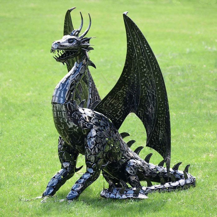 Dark metal dragon statue standing on grass with raised wings and a curled, spiked tail.