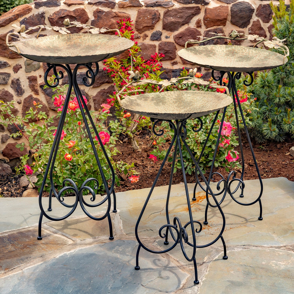 Three shallow birdbaths on ornate black stands arranged on a stone patio beside flowers.