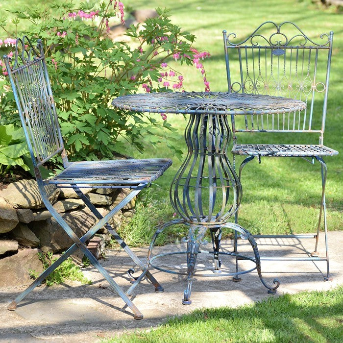Two ornate metal folding chairs flank a round bistro table on a garden patio.