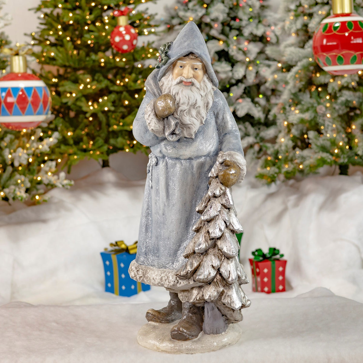 Full view of a blue-gray hooded Santa carrying a bag beside a snowy tree