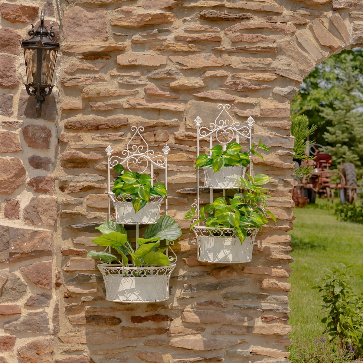 Two ornate white wall planters with leafy plants hang on a rustic stone wall beside a garden arch.