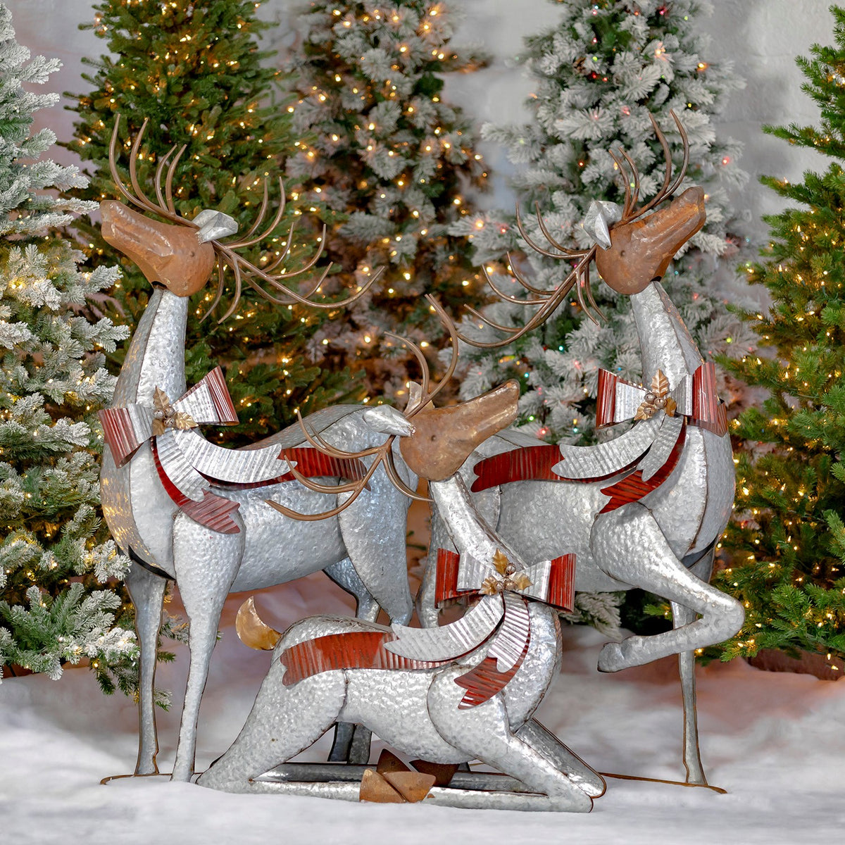 Three silver reindeer with brown antlers and red-and-silver bows pose among lit Christmas trees.