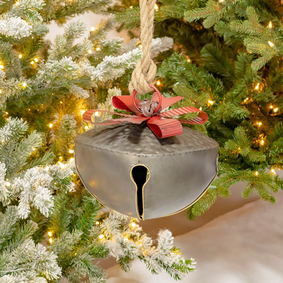 A broad galvanized bell with a red ribbon and rope hanger hangs among snowy, lighted evergreen branches.